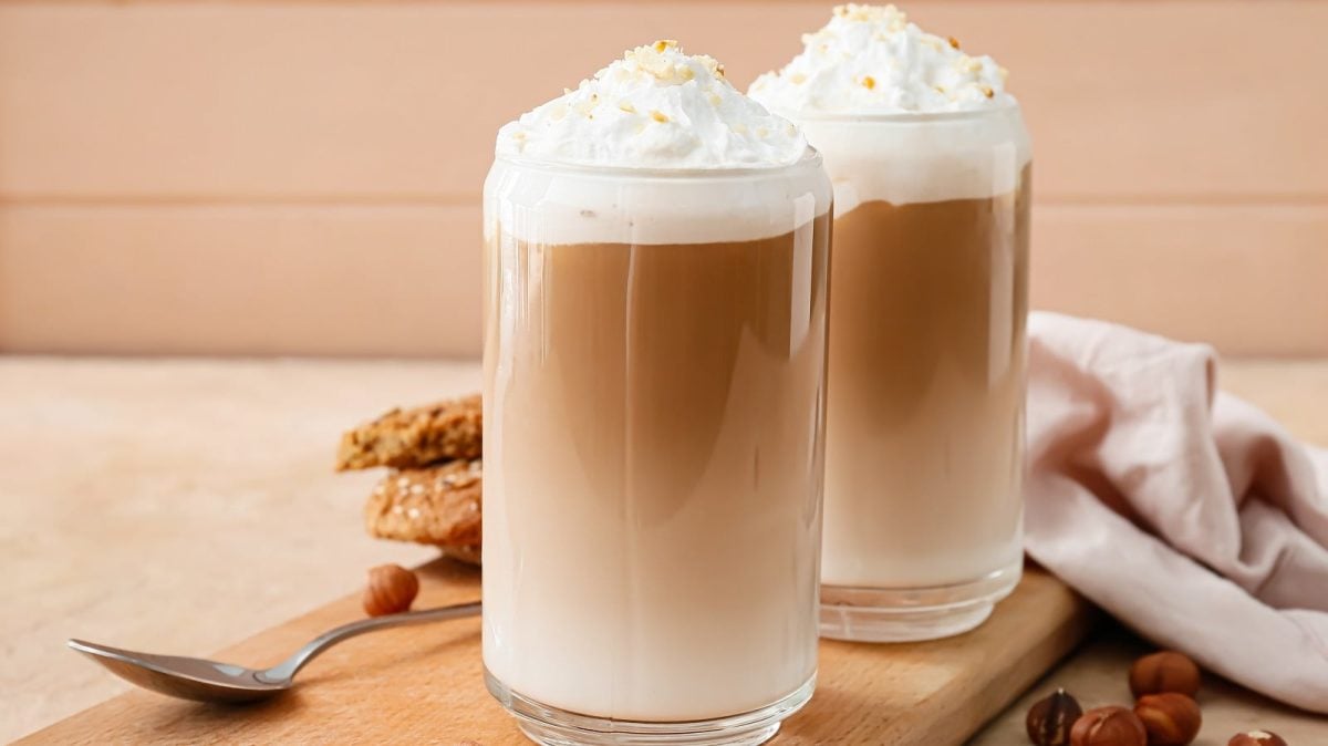 Zwei Gläser mit Haselnuss-Latte vor einem beigefarbenen Hintergrund.