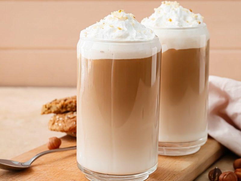 Zwei Gläser mit Haselnuss-Latte vor einem beigefarbenen Hintergrund.