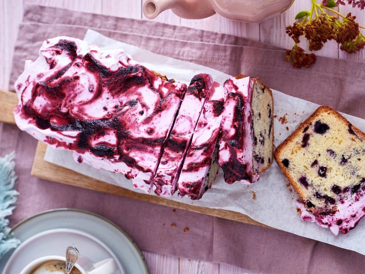 Ein Heidelbeerkuchen auf einem hölzernen Brett auf einem rosaroten Geschirrtuch auf einem rosa Hintergrund. Links unten eine Kaffeetasse und ein blaues Geschirrtuch mit Dessertgabeln.