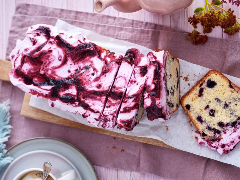 Ein Heidelbeerkuchen auf einem hölzernen Brett auf einem rosaroten Geschirrtuch auf einem rosa Hintergrund. Links unten eine Kaffeetasse und ein blaues Geschirrtuch mit Dessertgabeln.