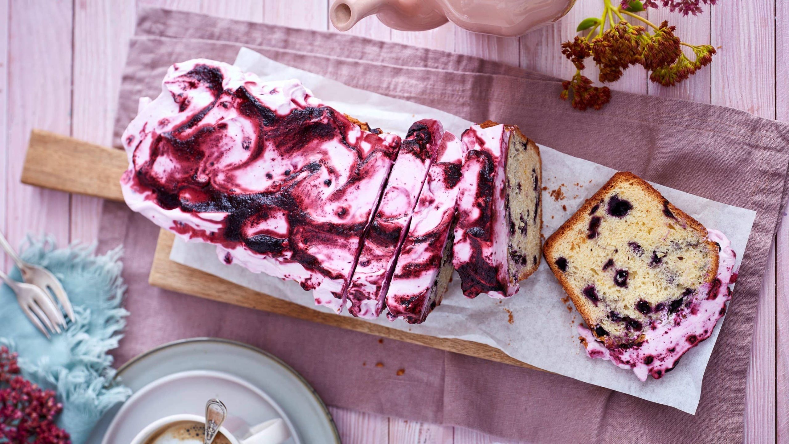 Ein Heidelbeerkuchen auf einem hölzernen Brett auf einem rosaroten Geschirrtuch auf einem rosa Hintergrund. Links unten eine Kaffeetasse und ein blaues Geschirrtuch mit Dessertgabeln.