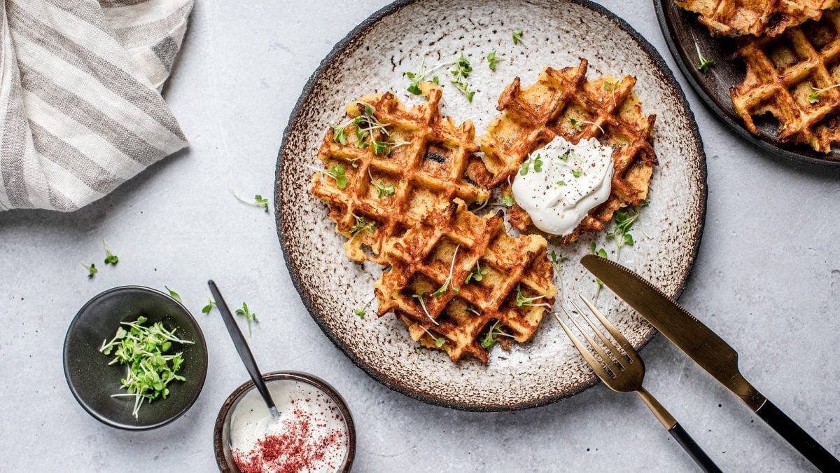 Ein Teller Reiswaffeln aus dem Waffeleisen, die mit einem Klecks Schmand bedeckt sind, daneben Messer und Gabel, ein Geschirrtuch, eine Schale mit Kräutern und Dip und ein Teller weiterer Waffeln.