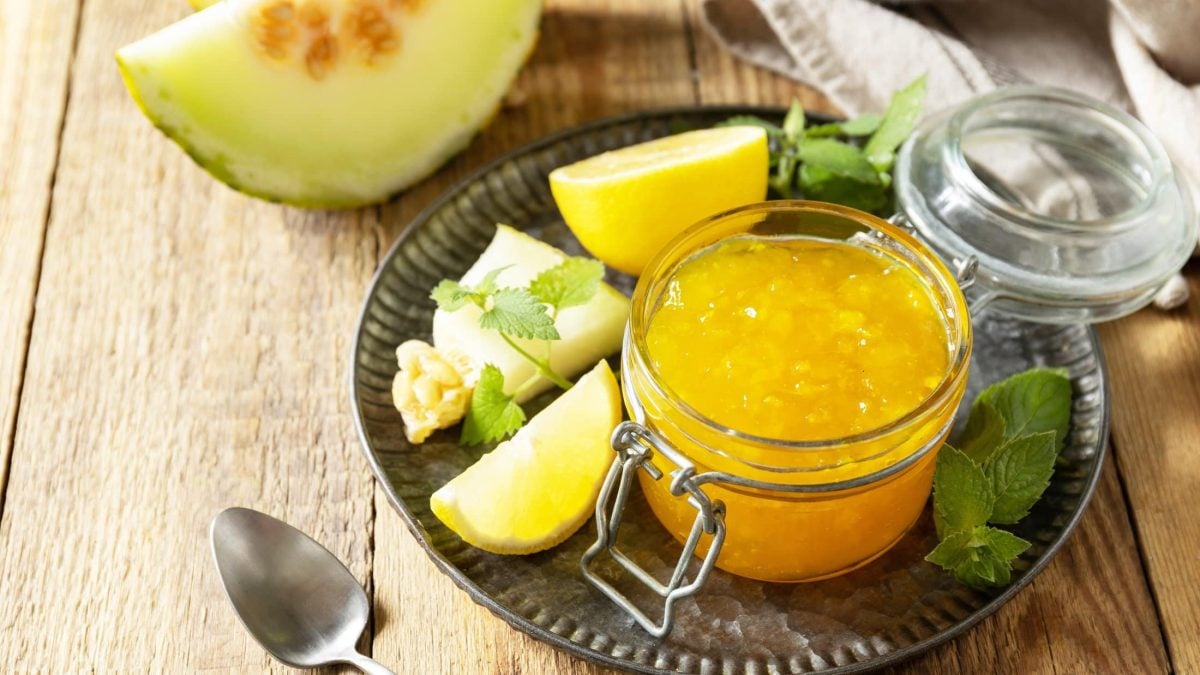 Draufsicht: ein Glas Honigmelonen-Marmelade auf einem dunklen Tablett. Drum herum liegen Honigmelonen-Stücke und frische Kräuter.