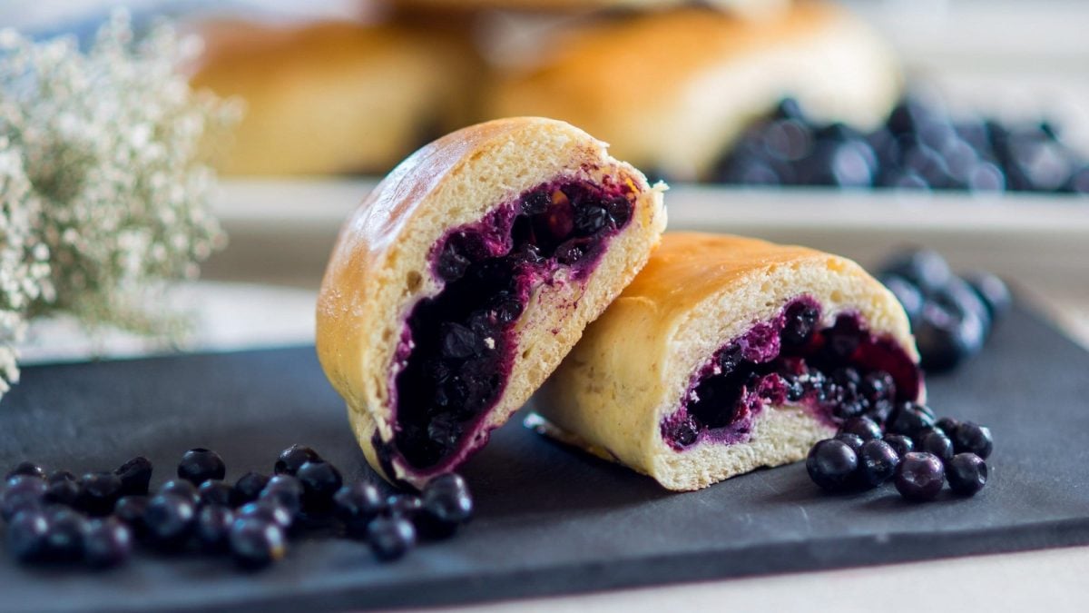 zwei angeschnittene Jagodzianki, polnische Hefebrötchen mit Blaubeerfüllung, auf einer Schieferplatte, drumherum weiße Blüten und frische Blaubeeren.
