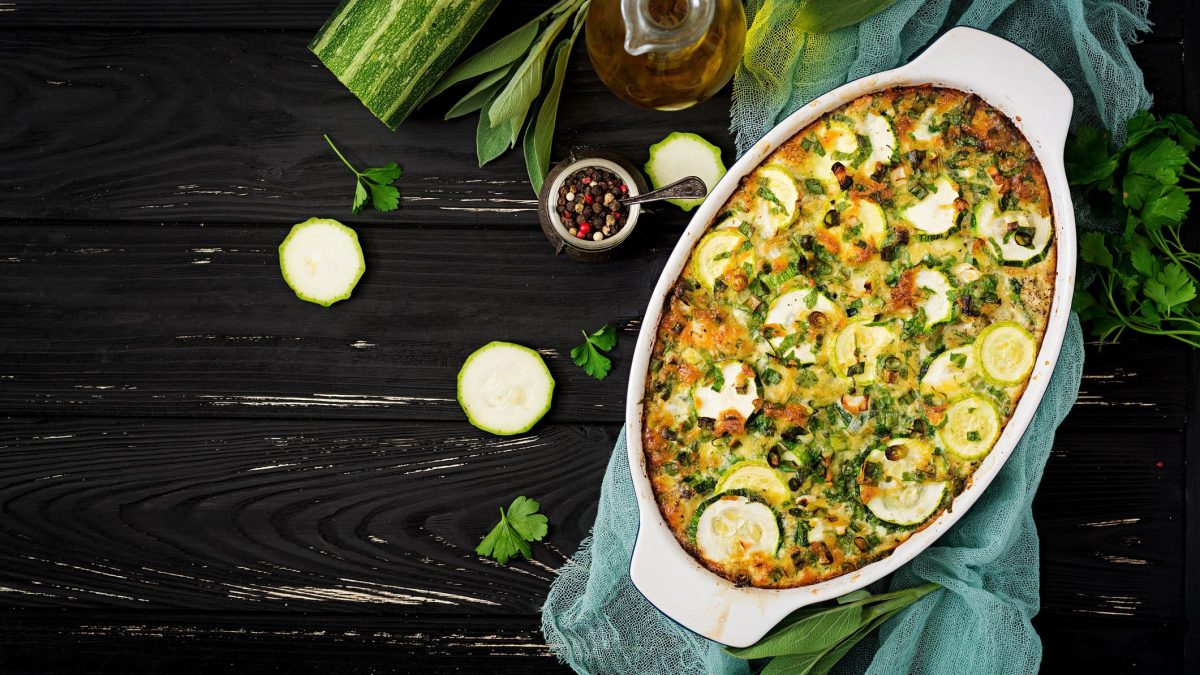 Kartoffel-Lauch-Auflauf mit Zucchini in weißer Auflaufform auf dunklem Holz. Daneben liegen verschiedene Zutaten.