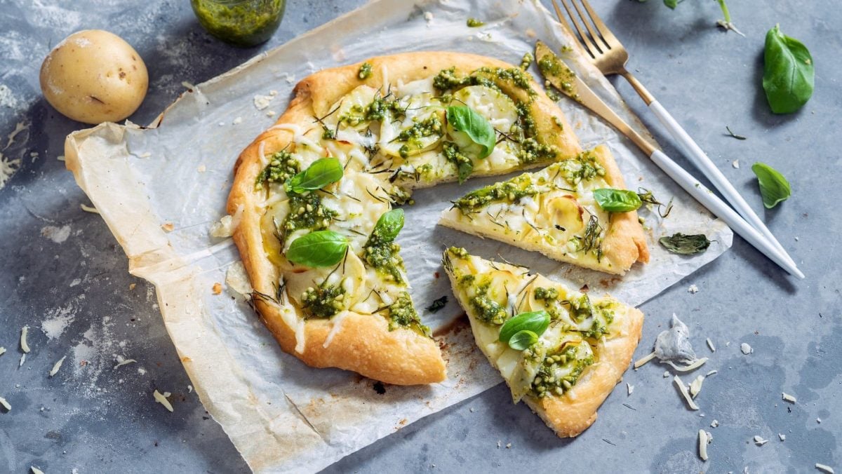 Draufsicht: eine angeschnittene Pizza di Patate liegt auf Backpapier auf einem Backblech. Auf dem blau-grauen Untergrund liegen noch eine Kartoffel, Besteck und frische Basilikumblätter.