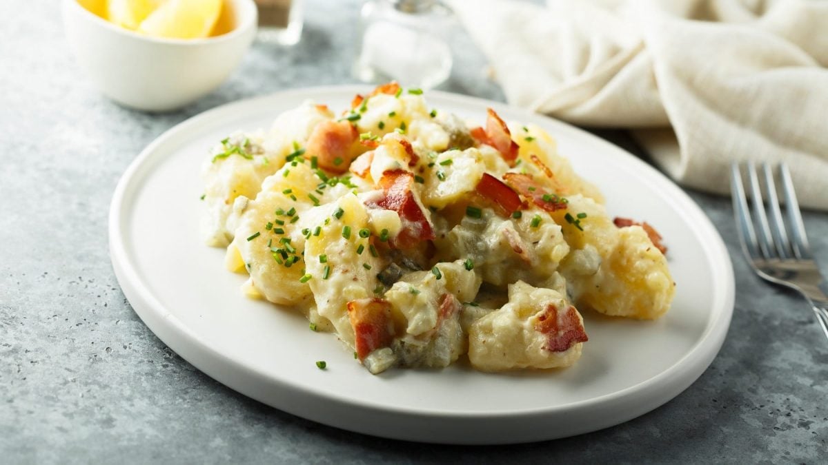 Ein weißer Teller mit dem Kartoffelsalat mit Speck und Cornichons auf grauem Untergrund. Im Hintergrund eine helle Serviette sowie Salz- und Pfefferstreuer.