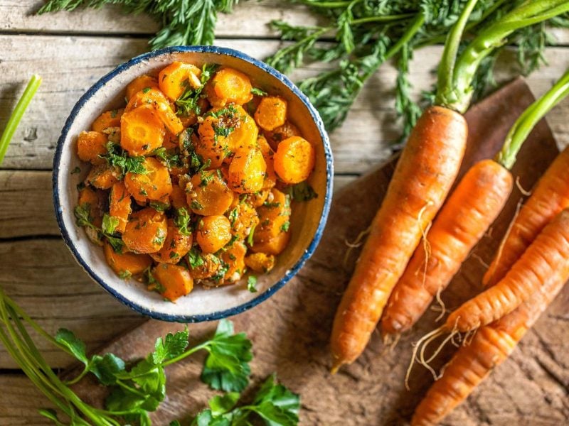 Eine Schüssel Karotten mit Knoblauch und Kreuzkümmel in der Draufsicht, daneben liegen Karotten mit Grün.