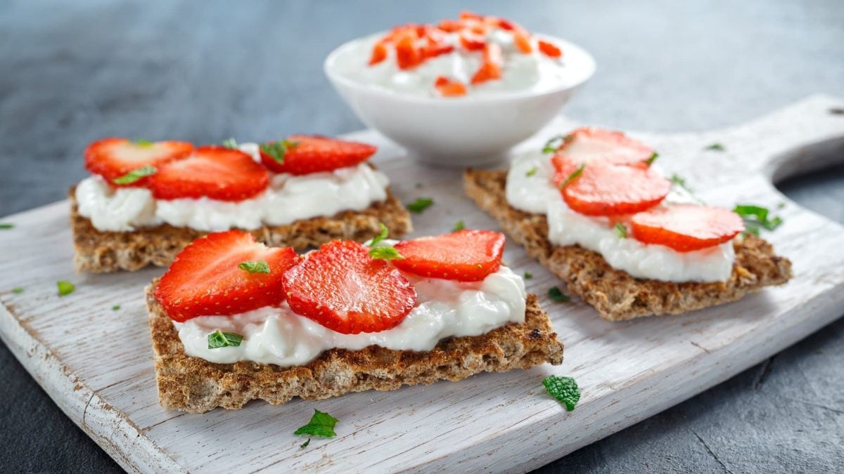 Frontal: Drei Knäcke mit Zitronen-Hüttenkäse und Erdbeeren liegen auf einem Holzbrett. Im Hintergrund ist eine schalte Hüttenkäse mit Errdbeeren.