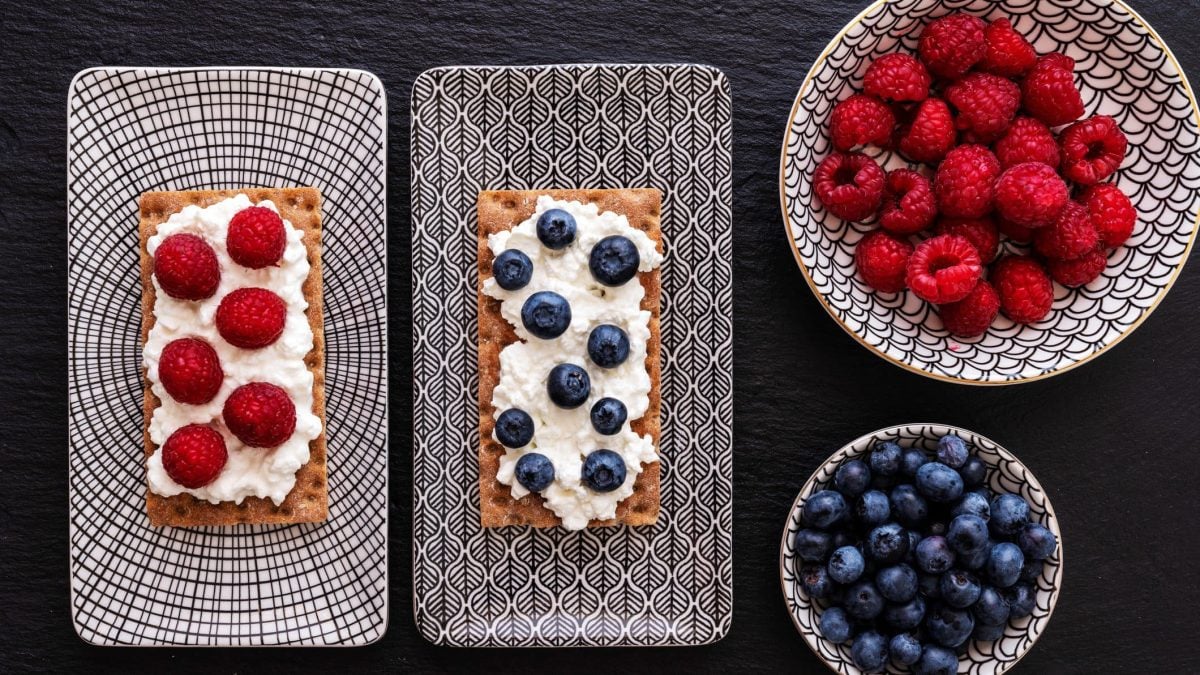 Zweimal Knäckebrot mit frischen Beeren und Vanillesaufstrich auf Frühstücksplatte auf dunklem Untergrund. Daneben zwei Schalen mit Beeren. Draufsicht.