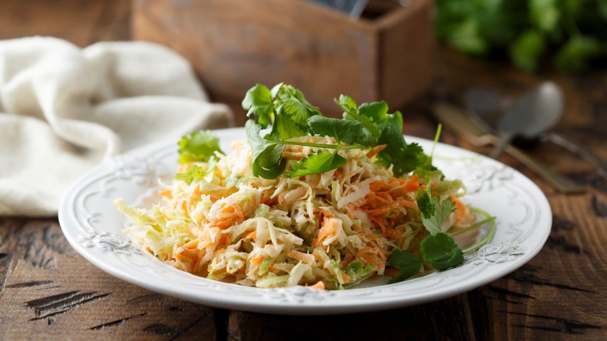Kohlrabi Slaw mit frischen Kräutern auf weißem Teller und Holzbrett. Im Hintergrund weißes Tuch, eine Holzschachtel und Kräuter. Frontalansicht.