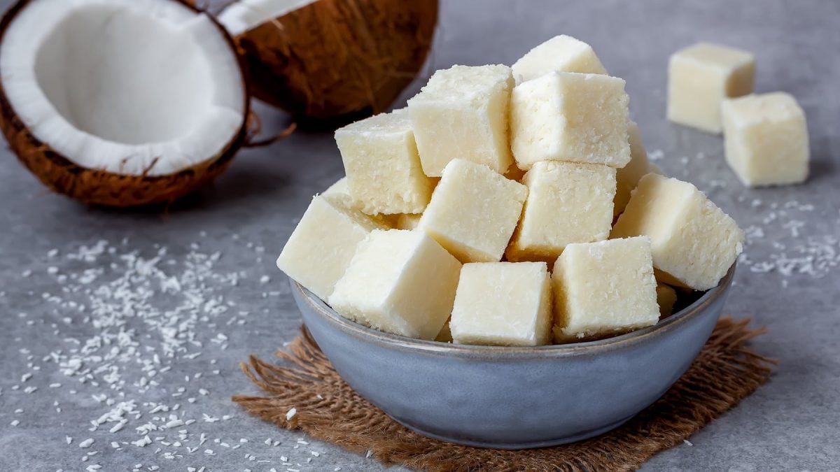 Weiße Kokos-Fudge-Würfel in einer blauen Schüssel auf einem grauejn Tisch mit frischer Kokosnuss und Koksraspel.