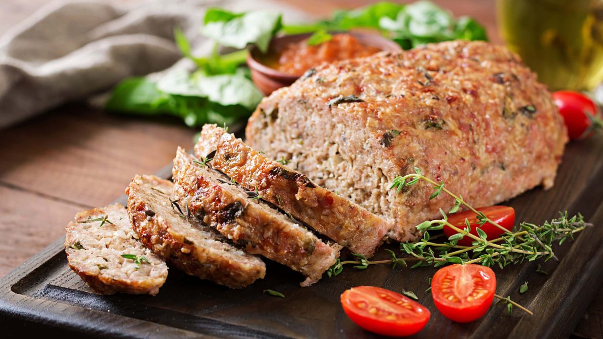 Ein Kräuter-Hackbraten auf einem dunklen Schneidebrett mit Tomaten und Thymian.