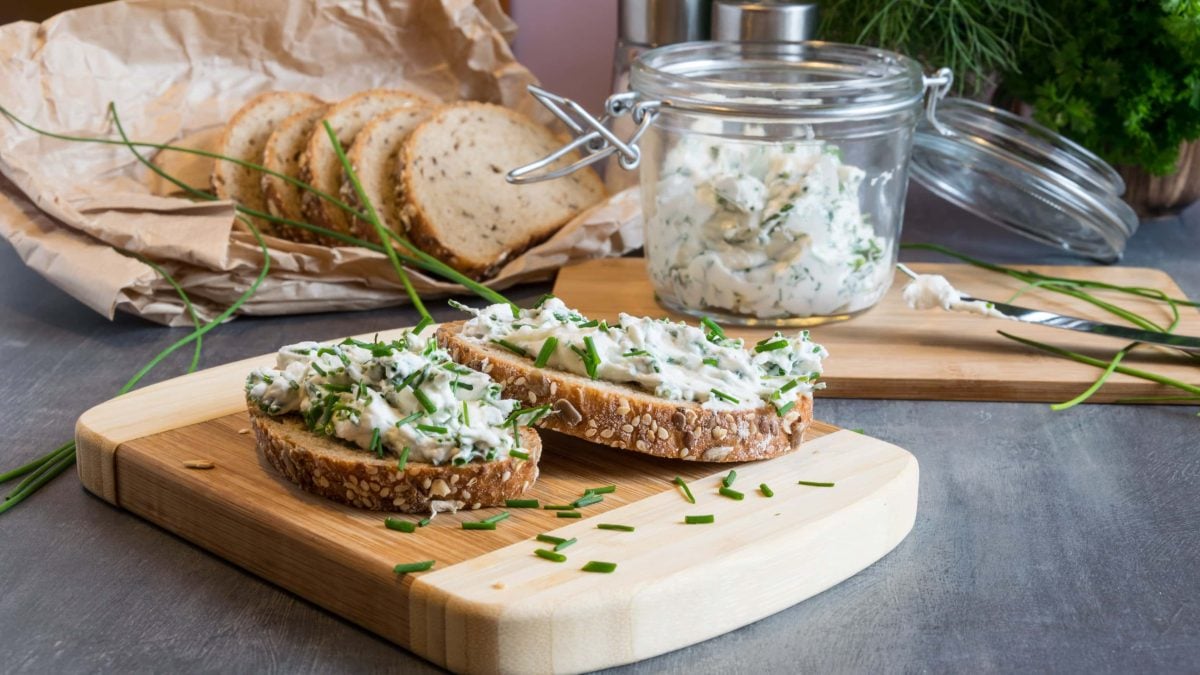 Frontal: Tisch, ein Brettchen mit zwei Scheiben Brot, die mit einem scharfen Kräuter-Hüttenkäse-Aufstrich bestrichen sind. Dahinter ein Glas Scharfer Kräuter-Hüttenkäse-Aufstrich, sowie weitere Brotscheiben. Drum herum liegt frischer Schnittlauch.