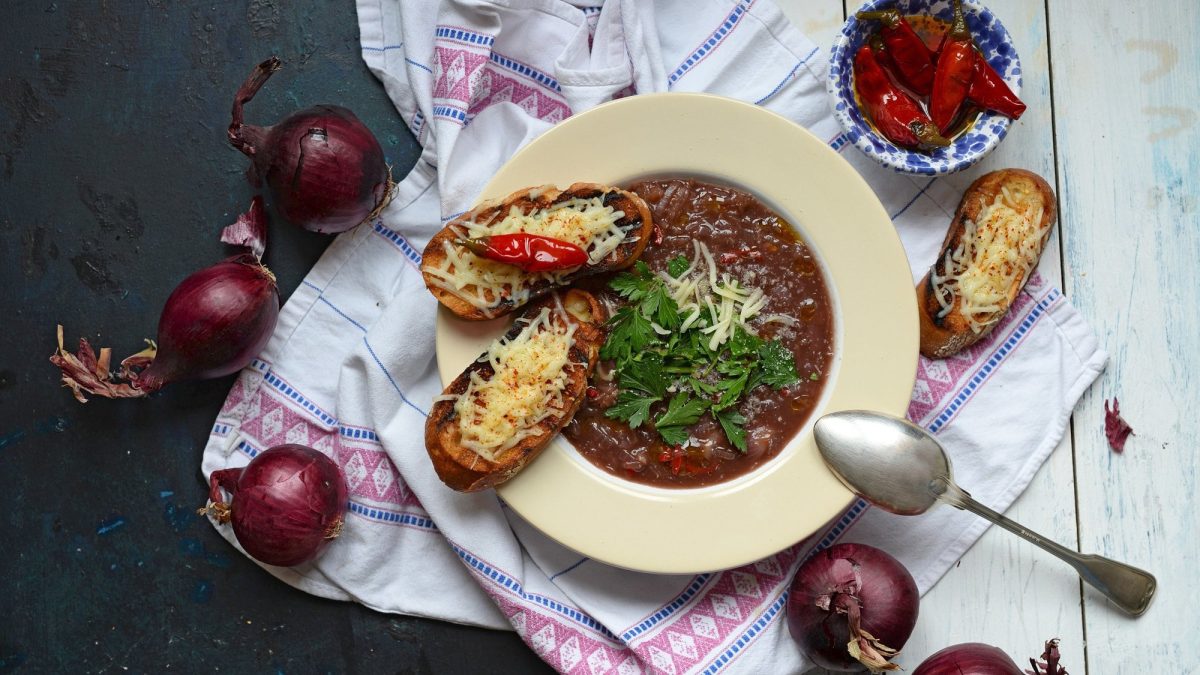 Ein Teller italienische Zwiebelsuppe Licurdia in der Draufsicht, darauf ein Löffel und geröstetes Weißbrot, daneben rote Zwiebeln.