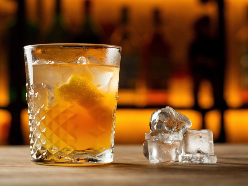 Ein Glas mit der Lynchburg Lemonade auf einem Holztresen. Im Hintergrund unscharf weitere Flaschen und Gläser im Regal zu erkennen.