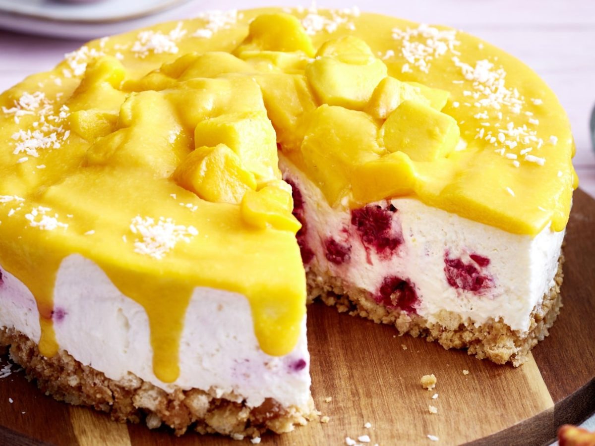 Eine Mango-Himbeer-Torte auf einem hölzernen Tortenteller. Rechts davon ein graues Geschirrtuch.
