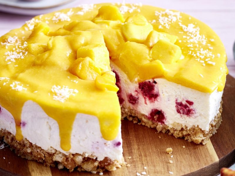 Eine Mango-Himbeer-Torte auf einem hölzernen Tortenteller. Rechts davon ein graues Geschirrtuch.