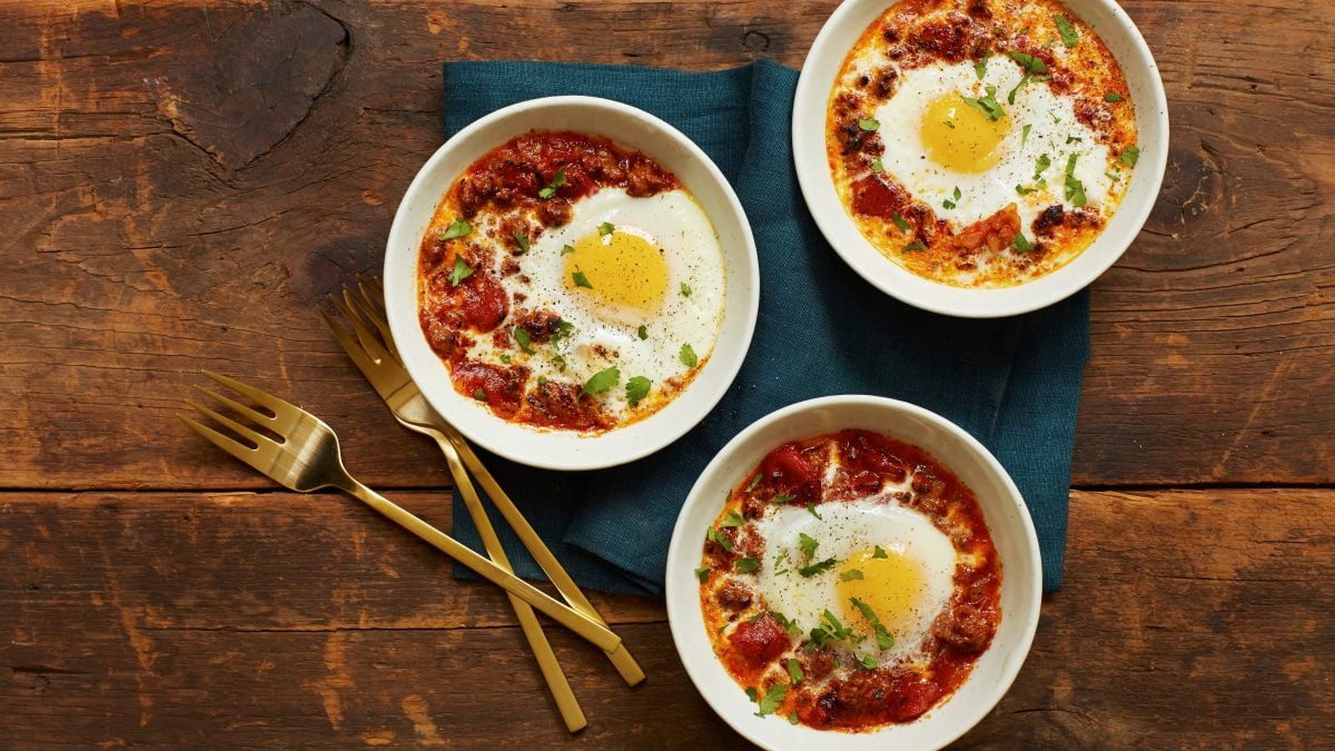 Merguez-Tajine mit Spiegelei und Ziegenfrischkäse in drei weißen Schüsseln auf blauem Tuch und Holztisch. Im Bild goldene Löffel. Draufsicht.