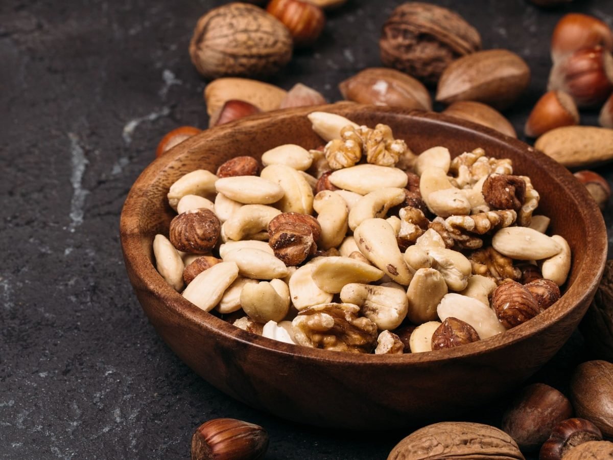 Nüsse gesund: Eine braune Holzschale mit einem Nuss-Mix seitlich auf schwarzem Marmor-Untergrund fotografiert.