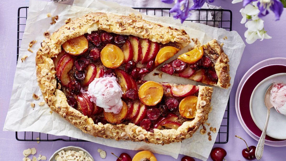 Eine angeschnittene Obst-Galette mit einer Kugel Eiscreme auf Backpapier, darunter ein schwarzes Gitter auf lila Untergrund, drum herum Früchte und Geschirr.
