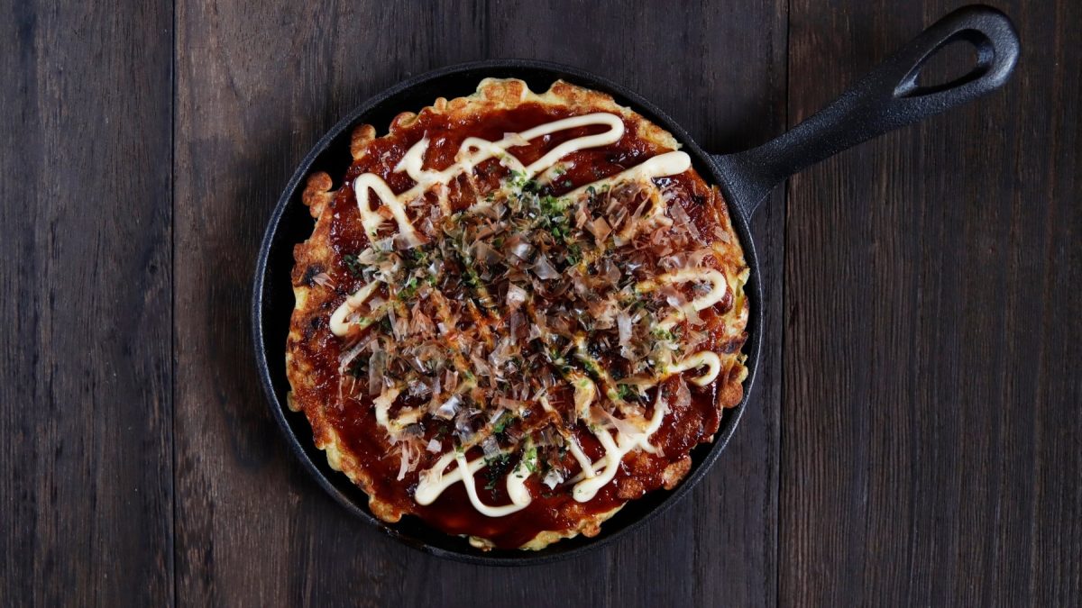 Eine schwarze Pfanne mit Okonomiyaki von oben fotografiert vor einem hölzernen Hintergrund.