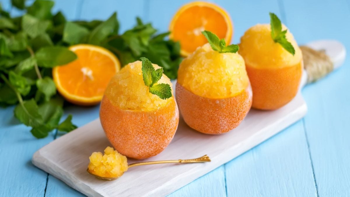 Drei ausgehöhlte Orangen mit dem Granita auf einem kleinen Schneidebrett und hellblauem Holztisch. Im Hintergrund Minze und Orangenhälften.