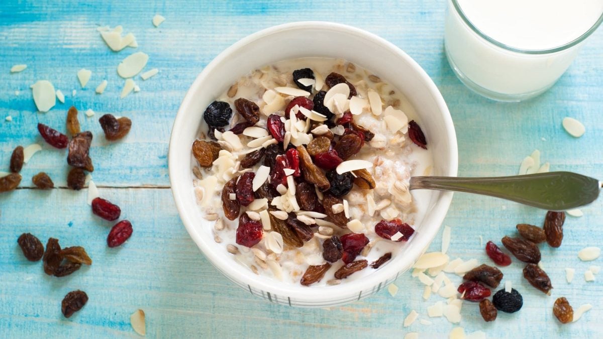 Hellblauer, melierter Untergrund aus Holz: Eine Bowl Overnight-Oats mit getrockneten Früchten und Mandeln steht darauf. Daneben steht ein Glas Milch. Auch Mandeln und getrocknete Früchte sind verstreut.