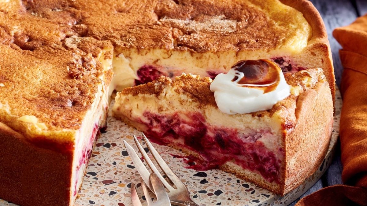 Ein angeschnittener Pfälzer-Zwetschgen-Cheesecake auf einem Servierteller. Rechts davon ein oranges Geschirrtuch.