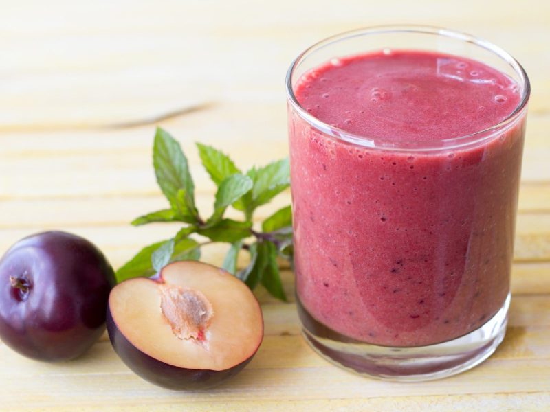 Pflaumen-Smoothie mit Heidelbeere, Minze und Basilikum in einem Glas auf Holztisch. Im Bild frische Minze und Pflaumen. Draufsicht.