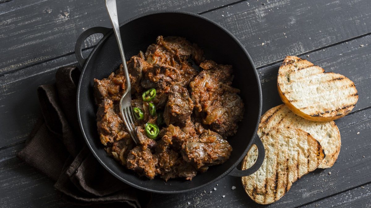 Draufsicht: Dunkler Untergrund aus Holz, eine dunkler offener Topf mit Piri-Piri-Hühnerleber und daneben liegen zwei geröstete Weißbrotscheiben.