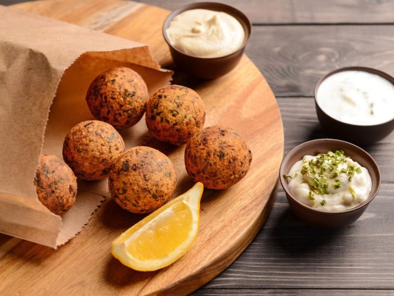 Popcorn Falafel aus Mais und Popcorn Kitchen Impossible in einer Papiertüre auf Holzbrett. Im Bild auf Holztisch dreierlei Dips und eine Zitronenspalte. Draufsicht.