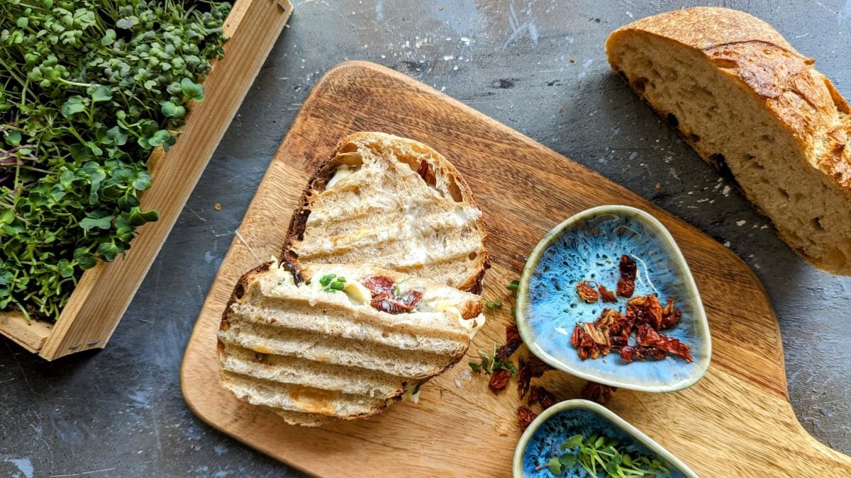 Das halbierte Sandwich auf einem Holzbrett. Daneben Kresse, Brot und getrocknete Tomaten.