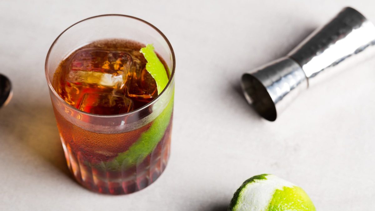 Rabo de Galo Cocktail im Glas mit Limettenzeste auf hellem Untergrund. Im Bild angeschälte Limette und Barmaß. Draufsicht.