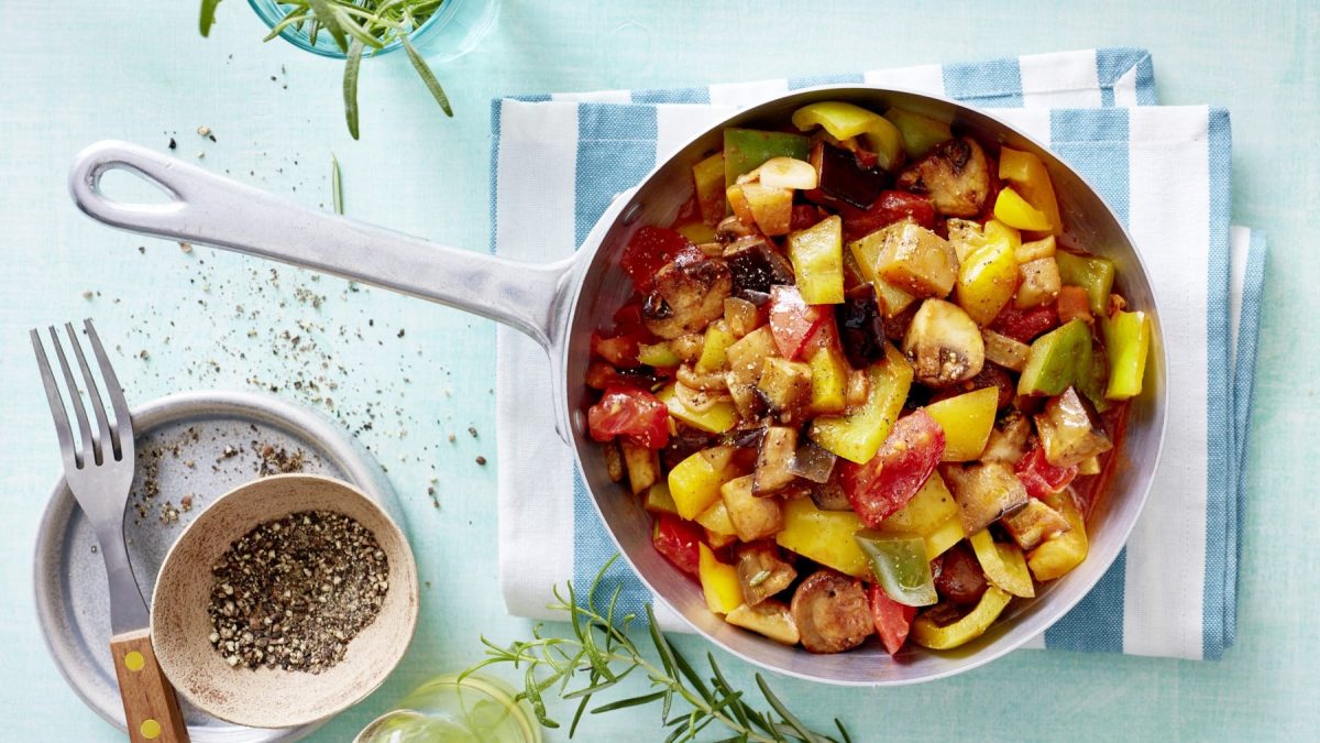 Eine Ratatouille-Pfanne auf einem blau-weiß gestreiften Küchentuch vor einem blauen Hintergrund. Links davon eine kleine Schüssel mit Pfeffer und eine Gabel. Oben und unten auf dem Bild Rosmarin.