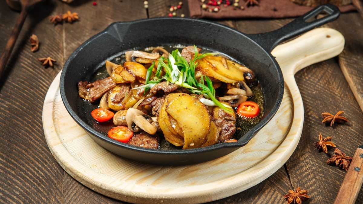 Eine gusseiserne Pfanne auf einem Holzbrett mit dem Rindfleisch-Kartoffelgericht auf dunklem Holzuntergrund mit Gewürzen. Seitlich von leicht schräg oben fotografiert.
