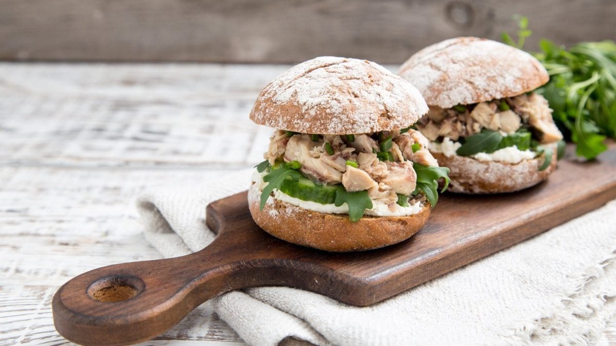 Zwei Roggenburger mit Hüttenkäse und Thunfisch Burger zum Frühstück auf Holzbrett mit Griff auf Tuch und hellem Untergrund. Im Bild Rucola. Frontalansicht.