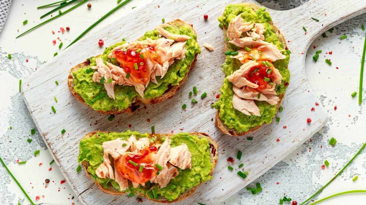 Drei belegte Brote mit Guacamole, Thunfisch und Sweet-Chili-Sauce von oben fotografiert. Alles auf einem hellen Holzbrett und hellem Holztisch.