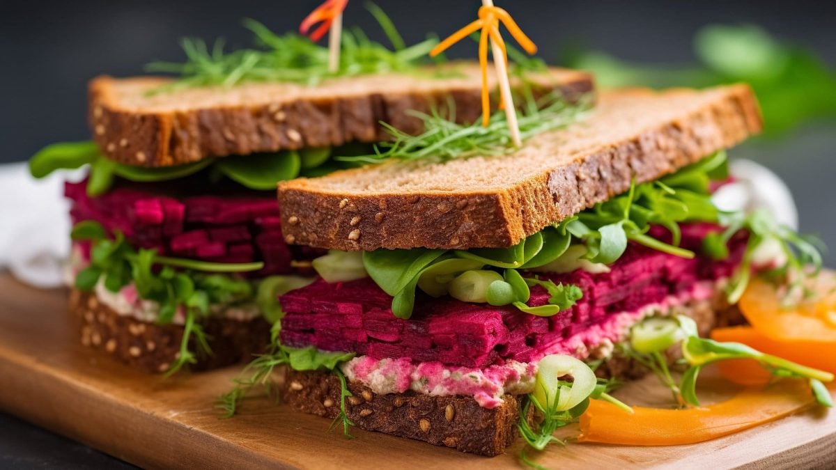 2 Sandwiches mit Rote-Bete-Hummus, Feldsalat, Frühlingszwiebeln und Frischkäse, fixiert mit einem Zahnstocher, auf einem Brett.