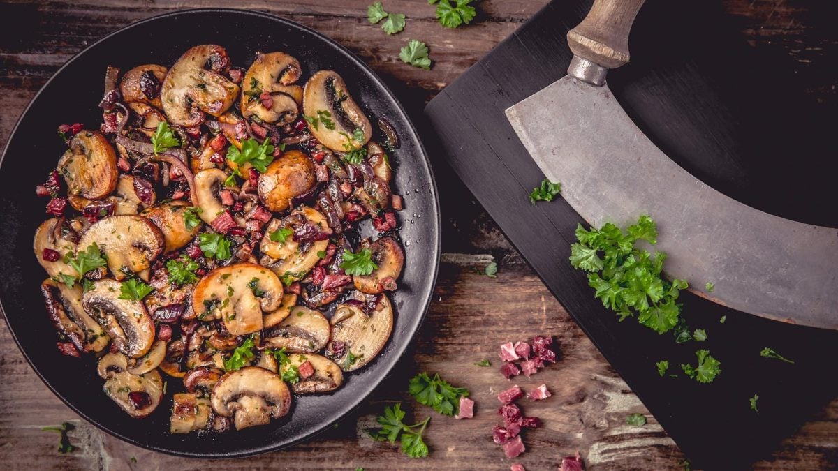 Draufsicht: eine Schinken-Champignon-Pfanne liegt auf einem Holztisch. Daneben liegen frisch gehackte Peterslilie und ein Wiegemesser,