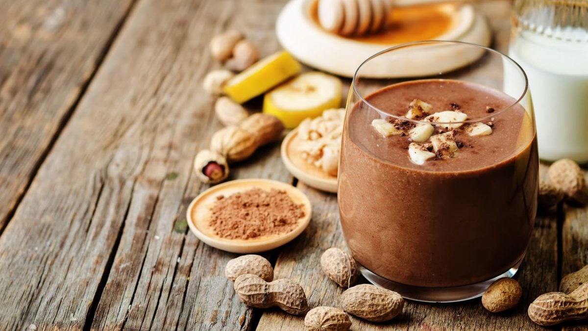 Ein Glas mit dem Shake sowie fast allen Zutaten, die reingehören (Nüsse, Banane, Kakaopulver) auf einem rustikalem Holztisch.