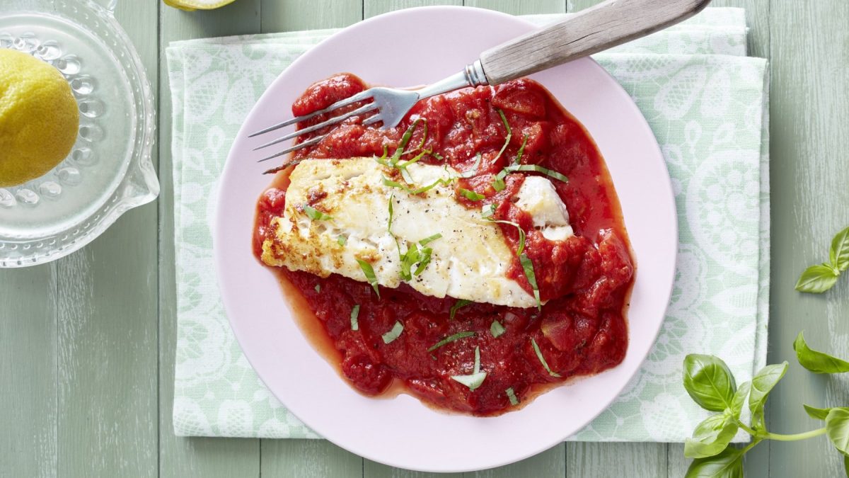Ein rosafarbener Teller mit Seelachs mit Tomatensugo auf einem hellgrünen Tisch.