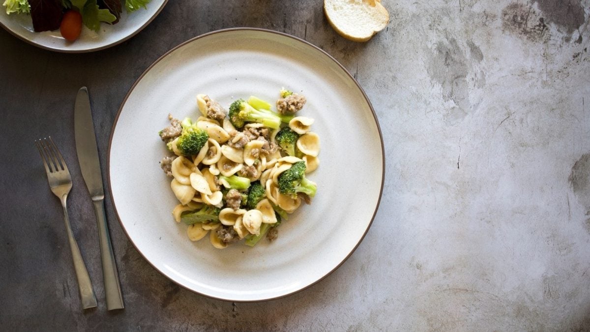 Ein weißer Teller mit Orecchiette mit Salsiccia und Brokkoli auf einem grauen Tisch.