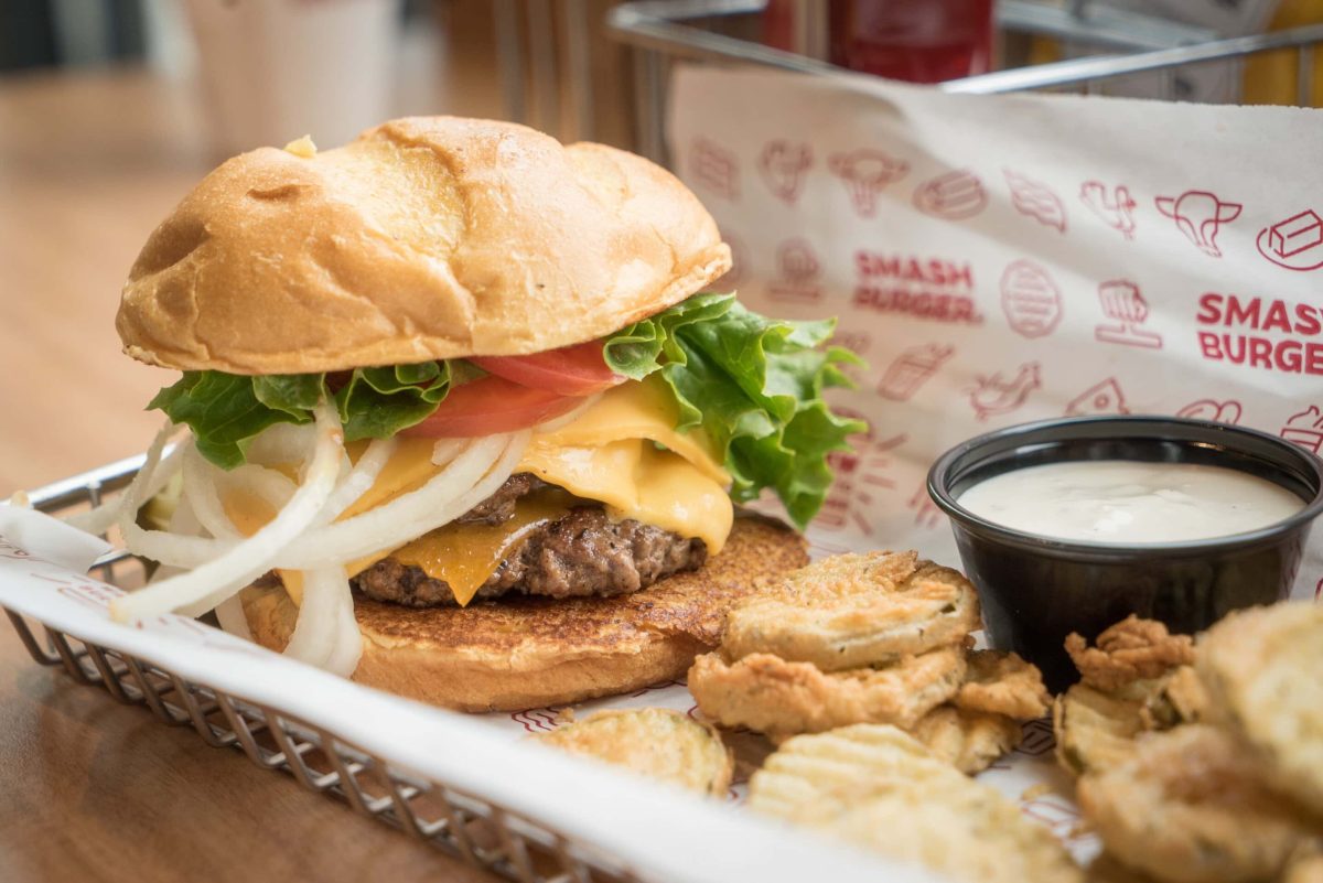 Was ist ein Smachburger? Unterschied zwischen Smashburger und normalem Burger. Smashburger mit frittiertem Gemüse und Dip auf weißer Servierplatte. Im Hintergrund eine Papier der Marke Smashburger. Frontalansicht.