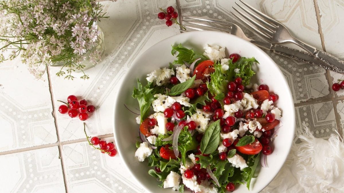 Draufsicht: Ein rot-weiß-kariertes Küchenhandtuch auf dem eine weiße Schüssel mit einem Sommersalat mit Johannisbeeren und Feta steht. Daneben ist einen Blumenstraß mit Holler zu sehen.