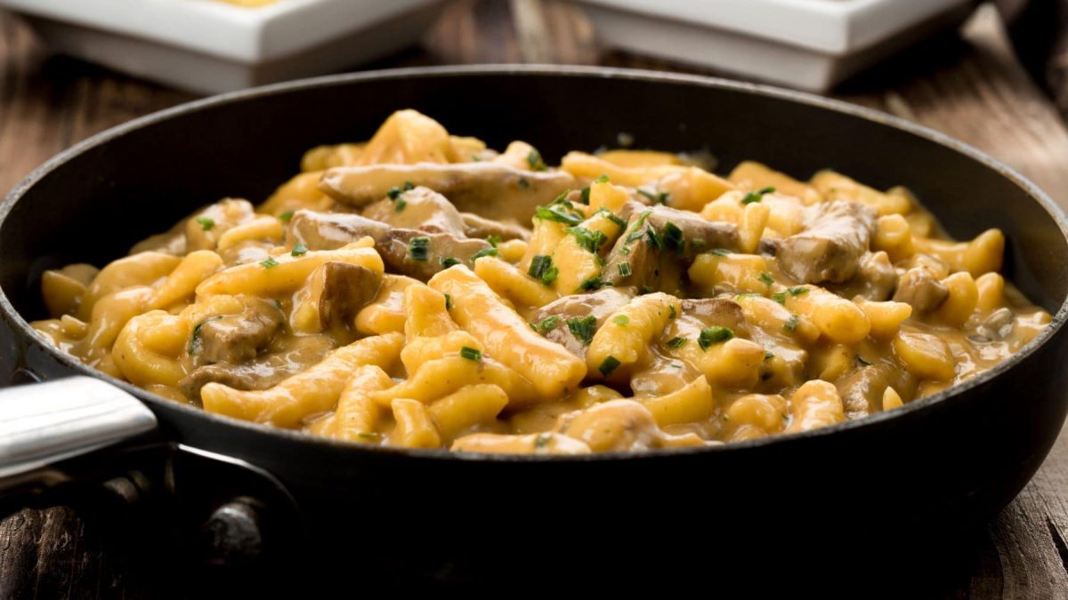 Spätzlepfanne mit Steinpilzen und Steakstreifen aus Flanksteak in einer schwarzen Pfanne auf Holztisch. Im Hintergrund angedeutet zwei weiße Schalen. Draufsicht.