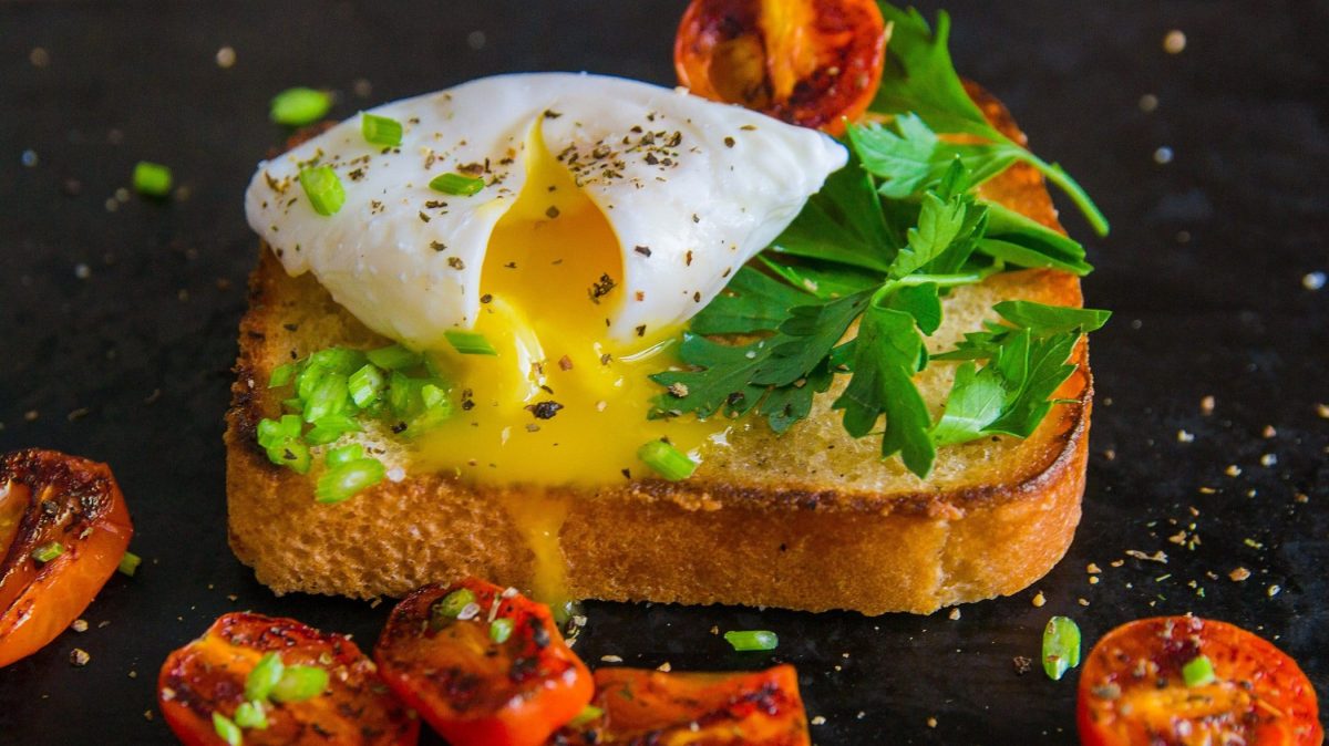 Frontalsicht: Auf einem dunklen Untergrund liegt ein Toast mit pochiertem Ei und gebratenen Tomaten und frischen Kräutern. Die Tomaten liegen um das Toast herum.