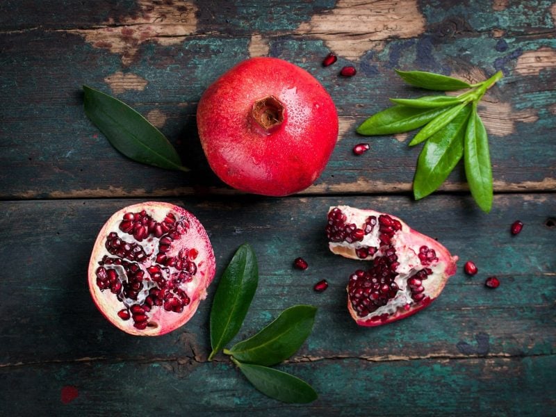 Granatapfel entkernen: ein geschlossener und ein geöffneter Granatapfel auf dunklem Holz mit grünen Blättern, Draufsicht.