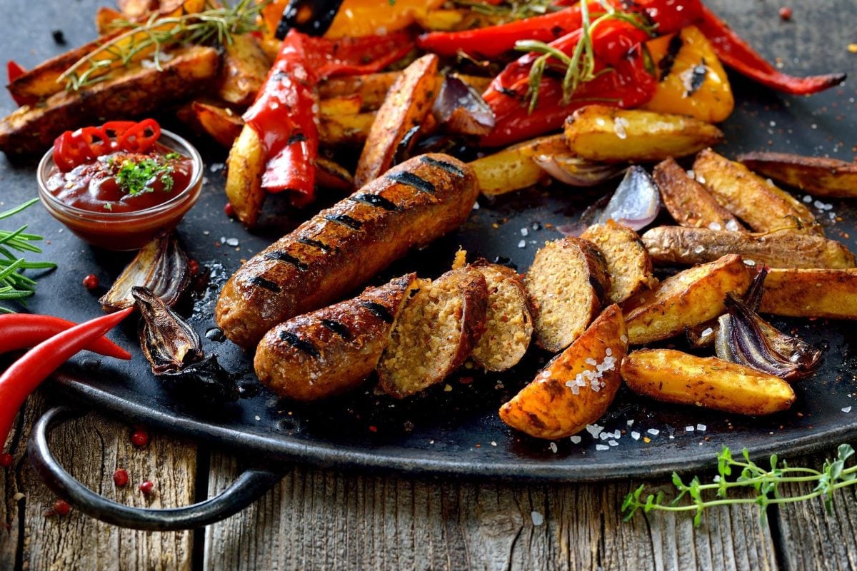 Vegane Bratwürste im Test: Zwei angeschnittene vegane Bratwürste auf einem Teller mit Kartoffelspalten und gegrillter Paprika.