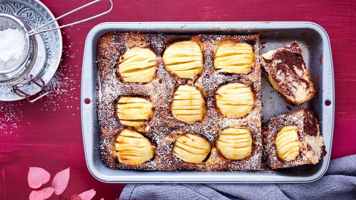 In einer Ofenform ist ein versunkener Apfel-Marmorkuchen gebacken. Er ist mit Puderzucker bestreut worden.